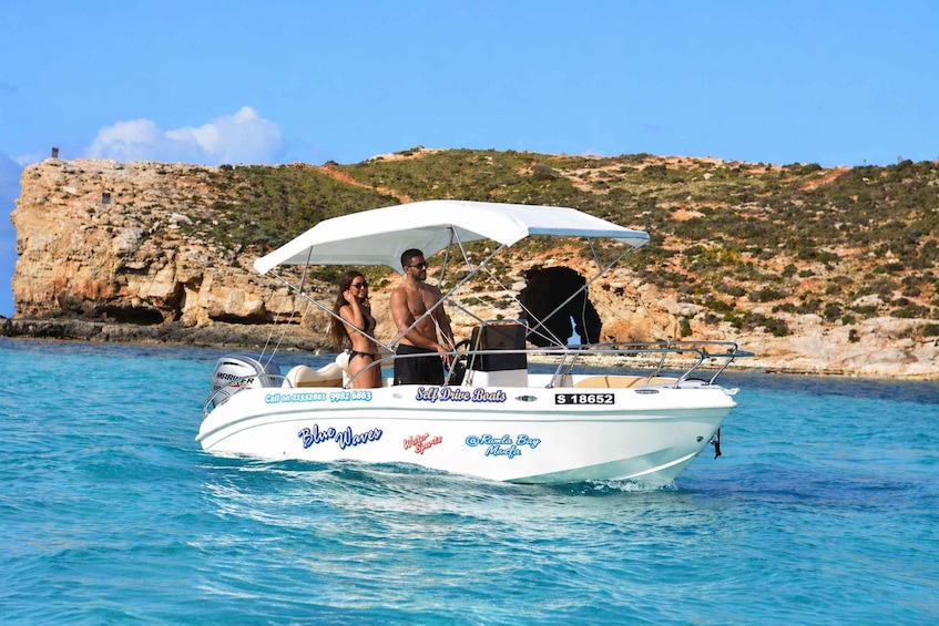 A white boat with 'Blue Waves' and 'Self Drive Boats' logos, a man and woman on board, and a rocky coastline in the background. 4