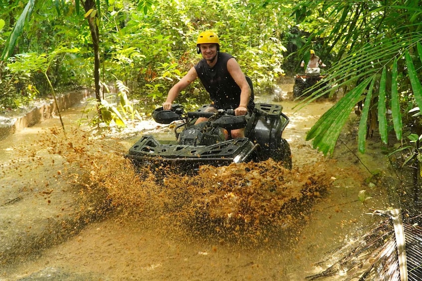 A person riding an ATV through a muddy area surrounded by dense greenery. 1