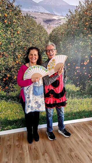 Two people holding fans in front of a backdrop with orange trees and a mountainous landscape. 2