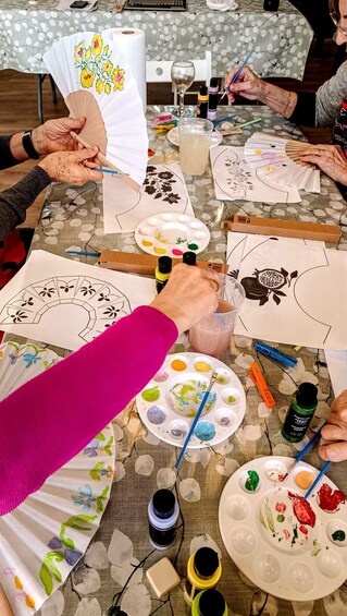 A group of people engaged in a painting activity, with various paint palettes, brushes, and paper fans on a table. 1