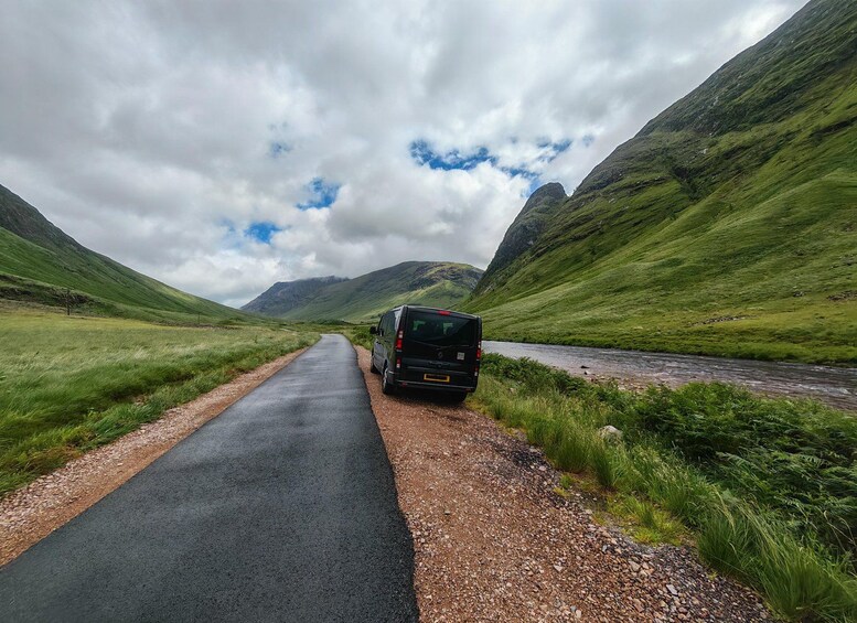 A black van parked on a gravel road beside a river, surrounded by green hills and a cloudy sky. 2