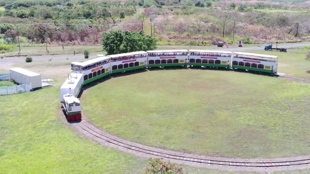 A train curving around a large open field, with a small building and a road in the background. 1