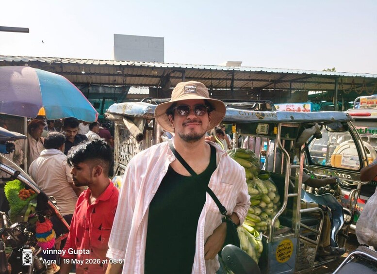 A person standing in a market with a hat, a green shirt, and a striped shirt over it. There are food stalls and a blue and white umbrella in the background. 3