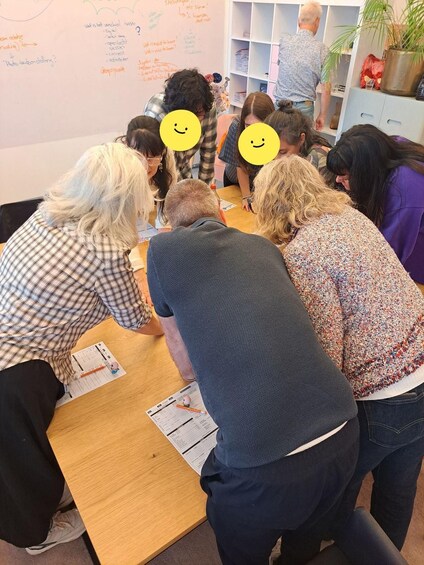 A group of people gathered around a table, engaged in a discussion or activity, with a whiteboard in the background. 1