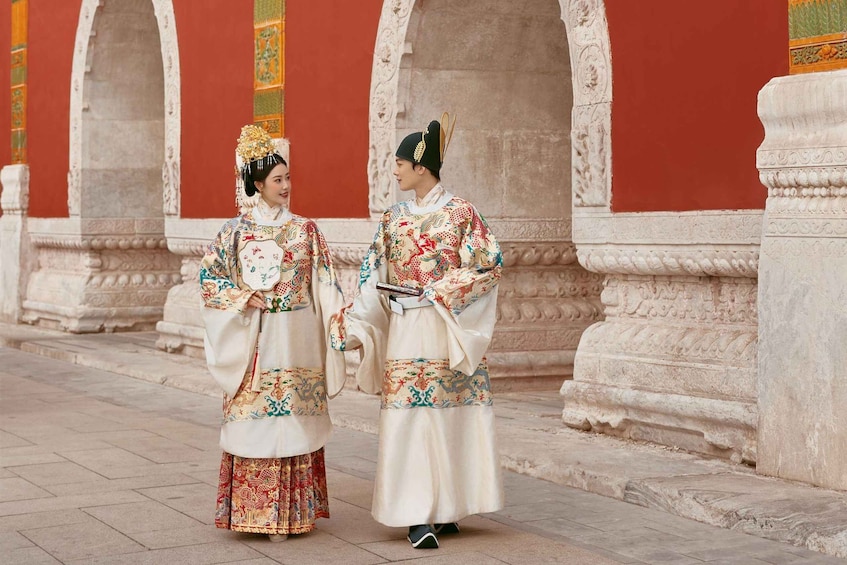 Two people in traditional attire walking in front of a red building with ornate columns. 1