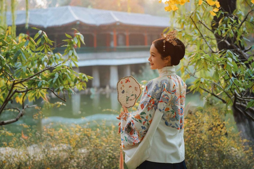 A person holding a decorative fan in a garden with a traditional building in the background. 4