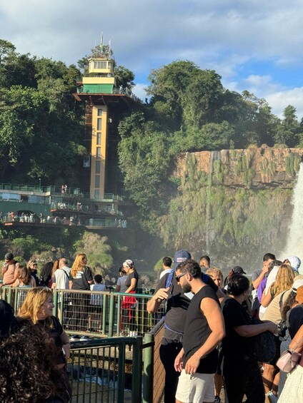 A crowd of people at a viewpoint overlooking a waterfall, with a tower structure in the background. 2