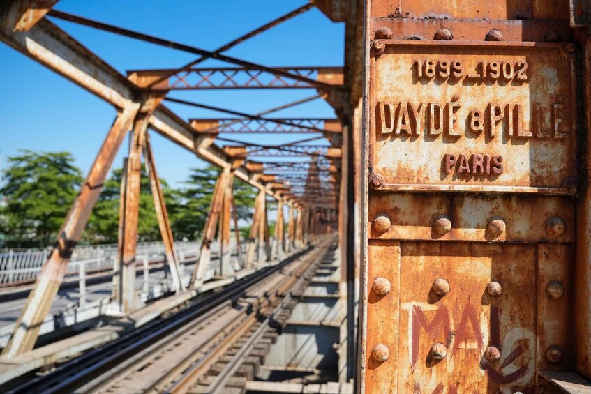 A rusty metal bridge with the inscription '1899-1902 DAYDE & PILE PARIS' and a railway track beneath it. 2