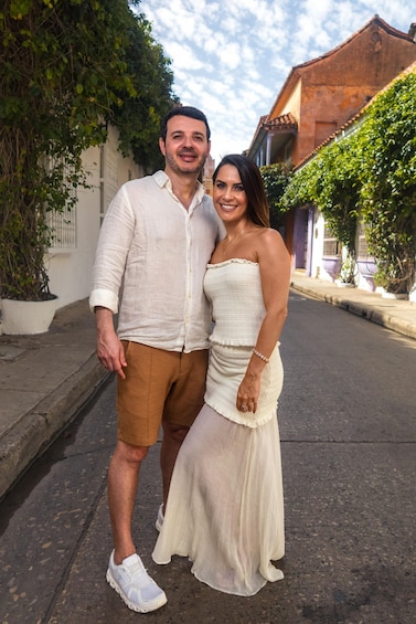 A couple standing on a street with buildings and greenery in the background. 3