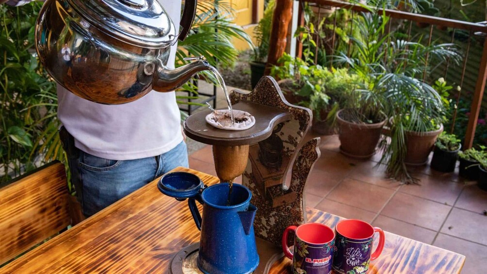 A person pouring coffee from a traditional metal coffee pot into a blue mug on a wooden table. 2