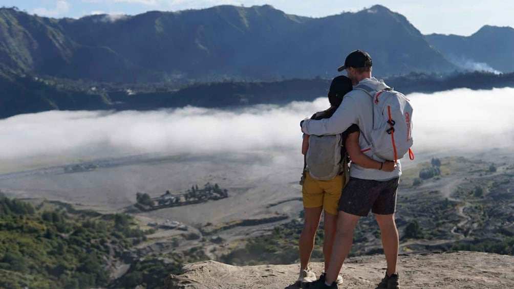 Two people standing on a mountain peak overlooking a valley with a misty forest below. 1