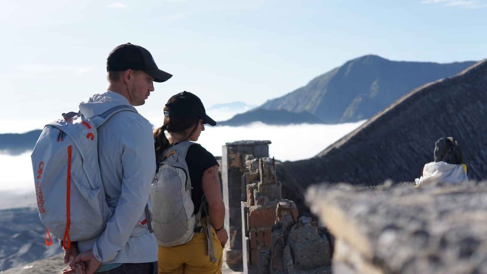 Two people standing on a mountain with a stone structure in the background and a mountain range in the distance. 5