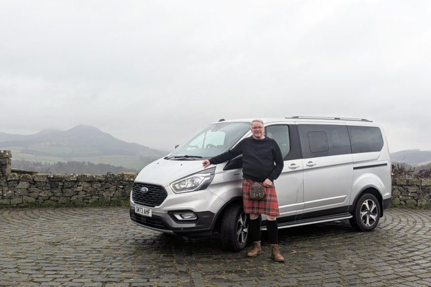 A person standing next to a silver minivan on a cobblestone road with stone walls and mountains in the background. 2