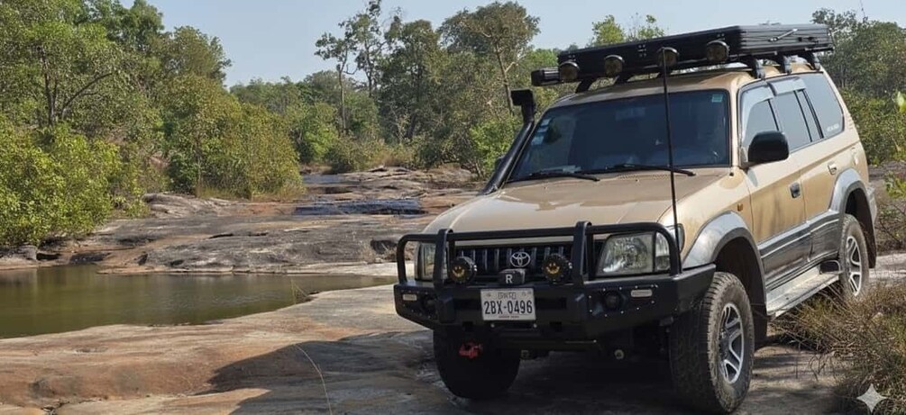 A beige SUV with a roof rack parked near a rocky riverbank. 5