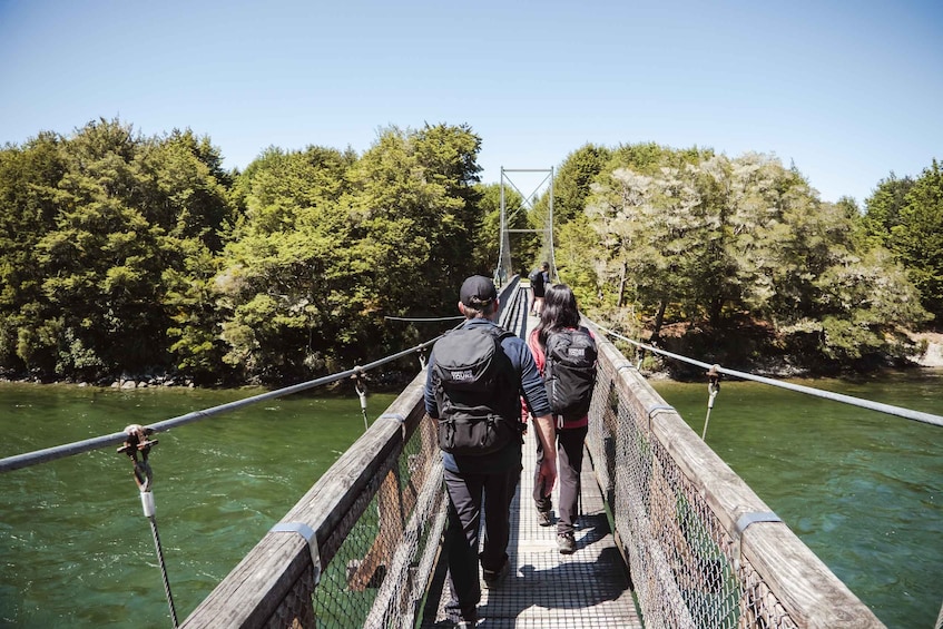 A group of people crossing a suspension bridge over a river, surrounded by dense forest. 1