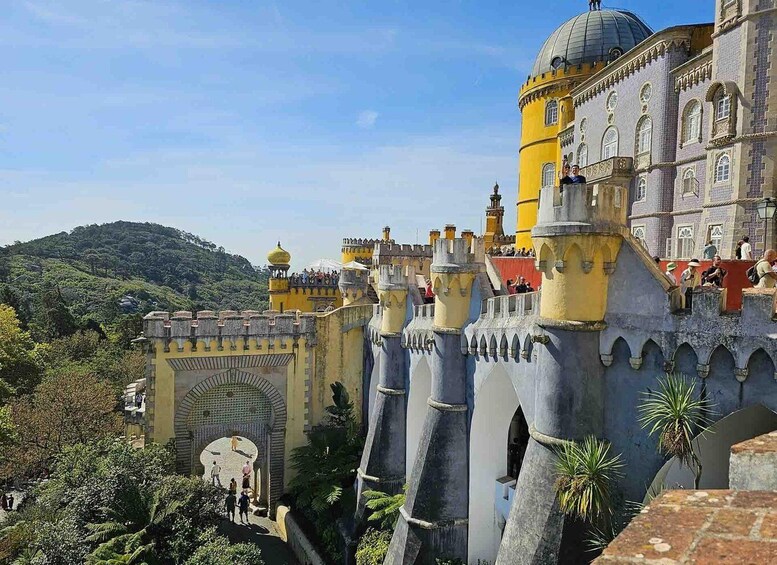 A castle with yellow and grey towers, surrounded by greenery and hills. 2