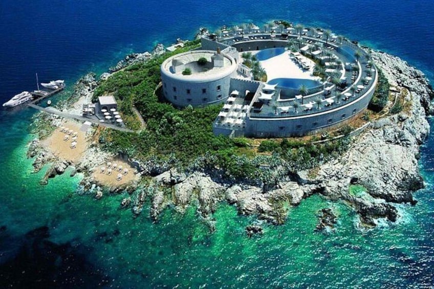 An island with a circular building surrounded by greenery and clear blue waters. 4