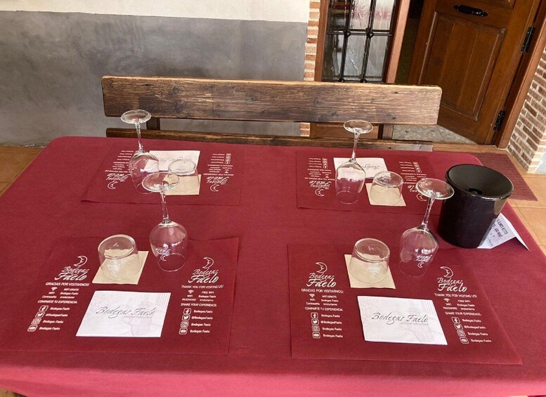 A table set with wine glasses and a black pitcher, covered by a red tablecloth with 'Bodega Faelo' printed on it. 4