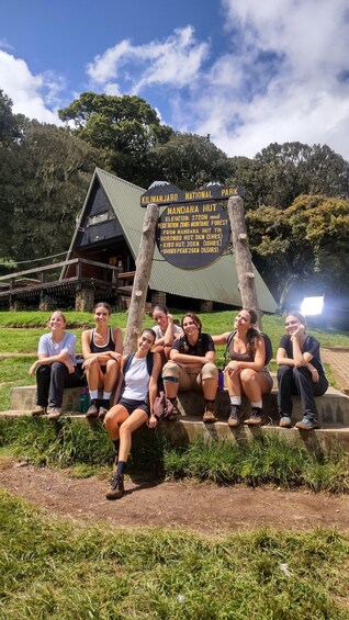 A group of people sitting on a concrete ledge in front of a sign for Kilimanjaro National Park, with a mountain and trees in the background. 4