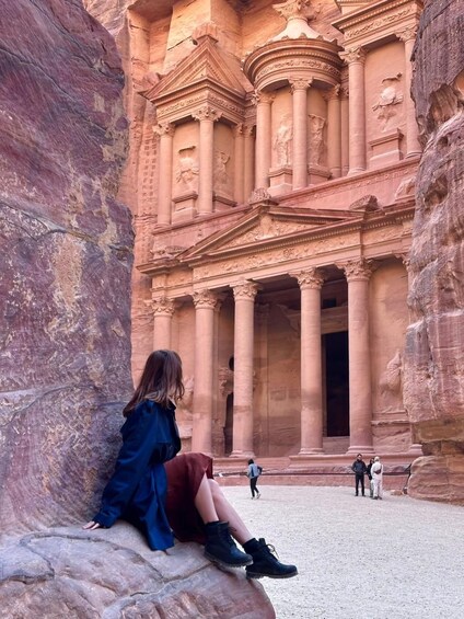 A person sitting on a rock in front of a large ancient structure with columns and detailed carvings. 4
