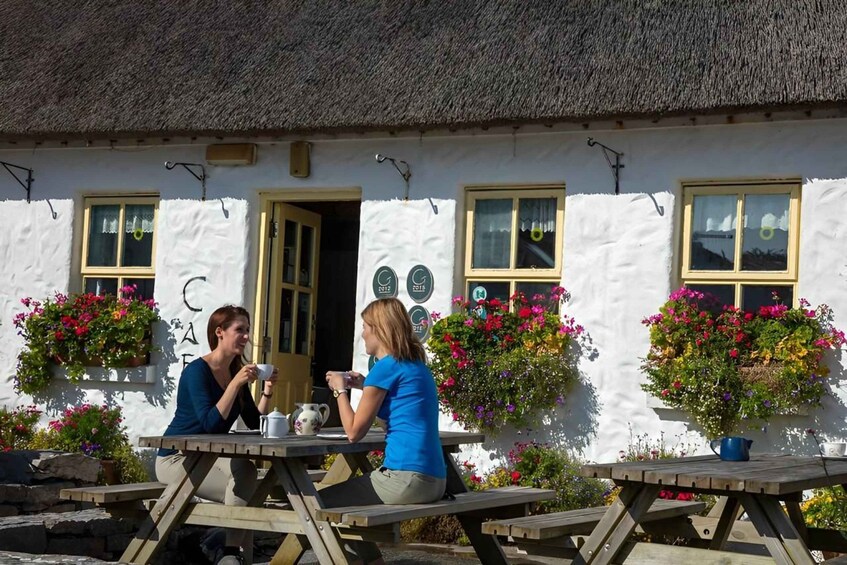 Two people sitting at wooden picnic tables outside a white building with a thatched roof, surrounded by colorful flowers. 3
