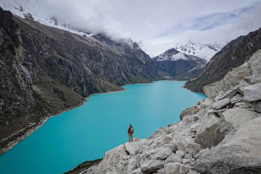 A person standing on a rocky outcrop overlooking a turquoise lake surrounded by mountains. 1