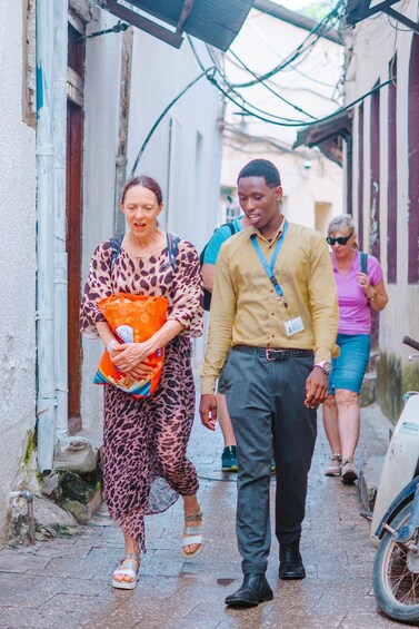 A narrow alley with wet pavement, a person in a leopard print dress holding a bag, and another in a yellow shirt walking beside them. 4