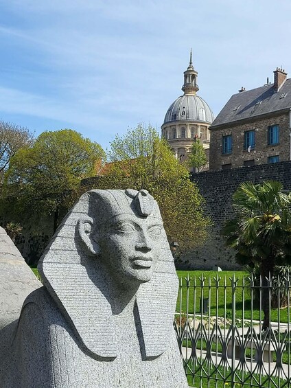 A stone sphinx statue in a park with a large building and trees in the background. 4
