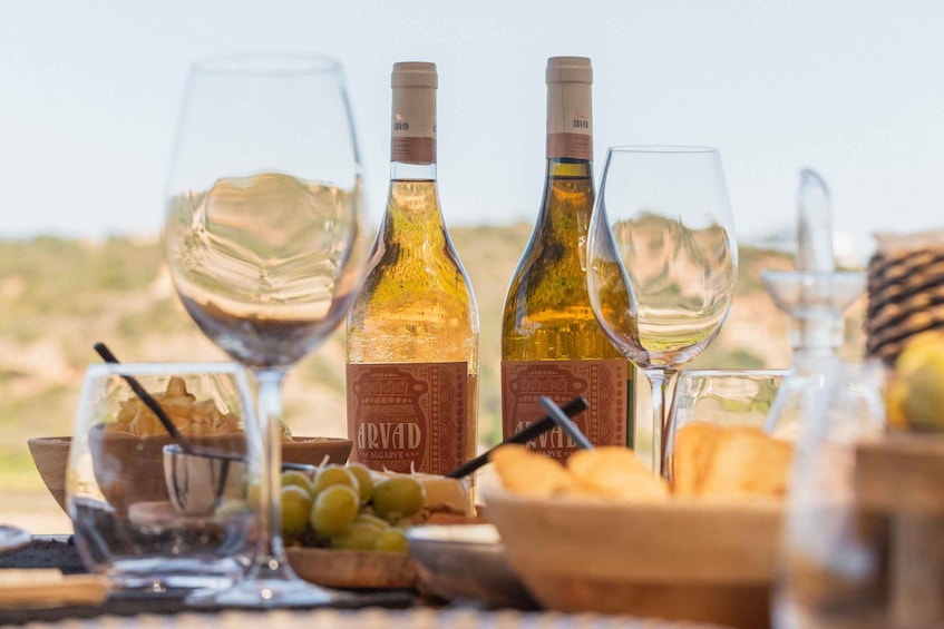 A table with two bottles of Arvad Alcarve wine, two wine glasses, a bowl of grapes, and a basket of bread. 1