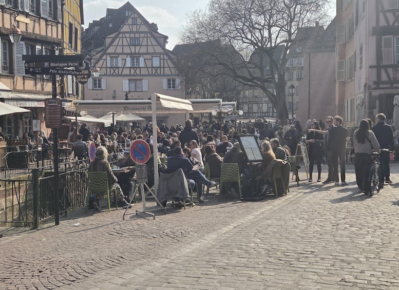 A crowded outdoor café with people sitting at tables, a no parking sign, and traditional half-timbered buildings in the background. 3