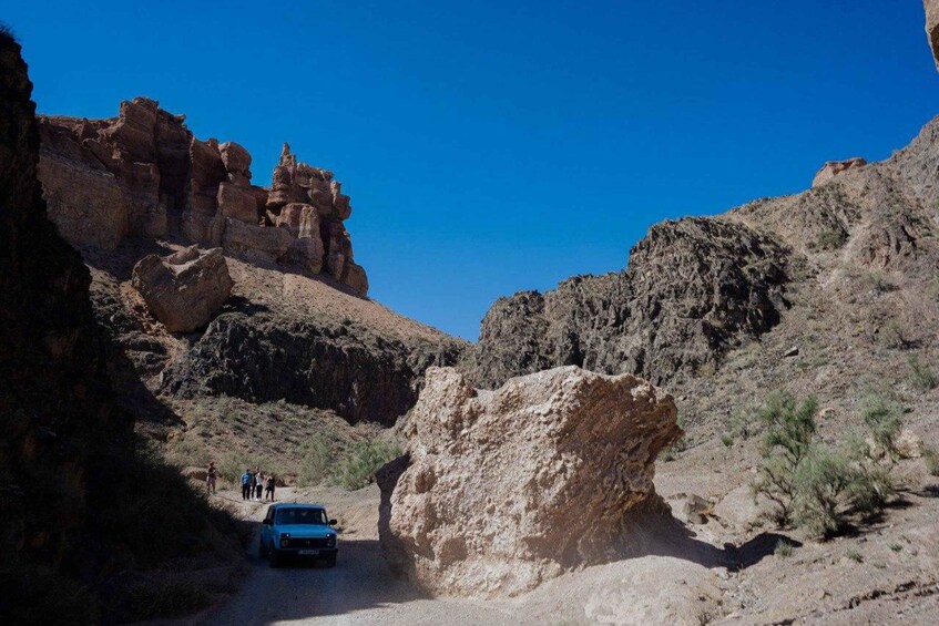 A rugged canyon landscape with a blue car navigating a narrow road, surrounded by rocky cliffs and sparse vegetation. 3