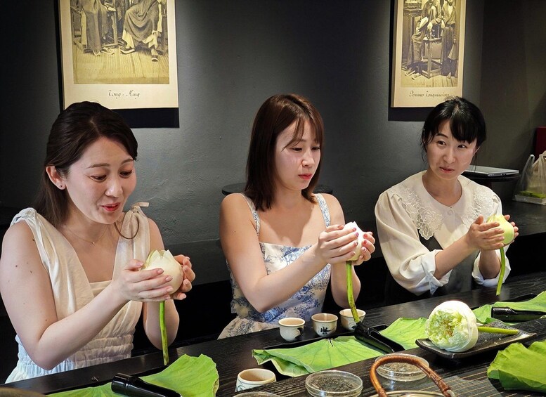 Three individuals engaged in a traditional food preparation activity, seated at a table with bowls and green leaves. 1