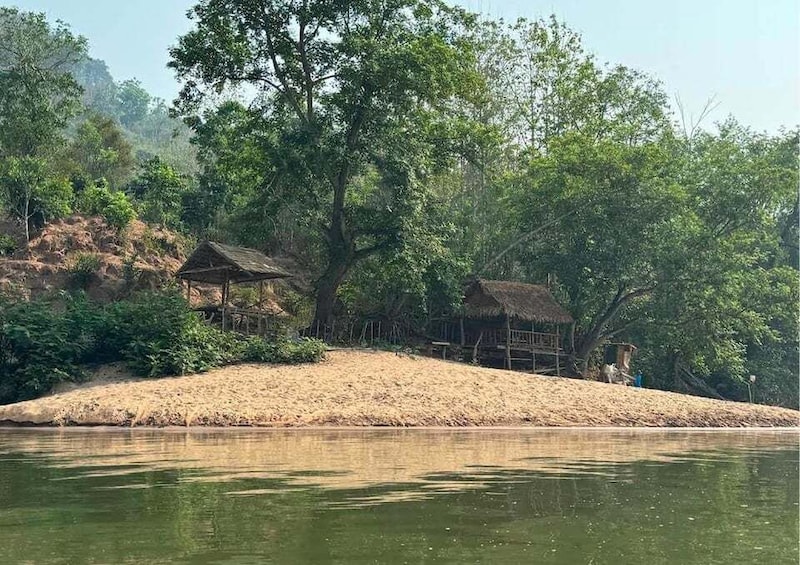 A riverbank with thatched-roof huts, dense greenery, and a sandy slope. 2