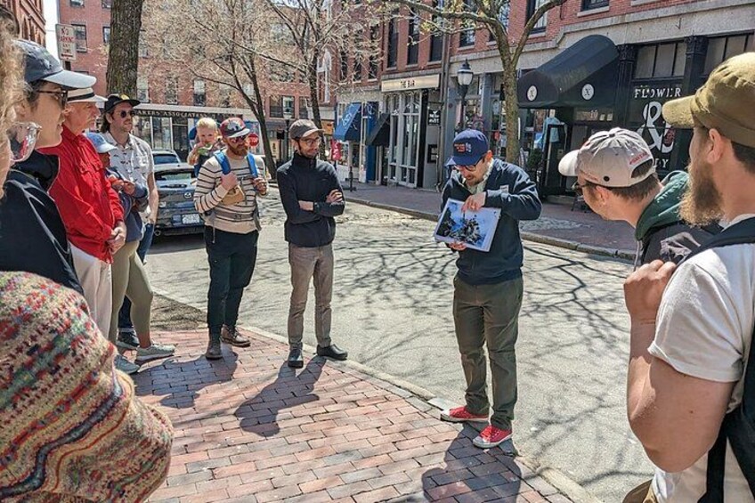 Un groupe de personnes se tient sur un trottoir, un homme tient une carte à la main, et un bâtiment porte une enseigne sur laquelle on peut lire « Fleurs ». 2