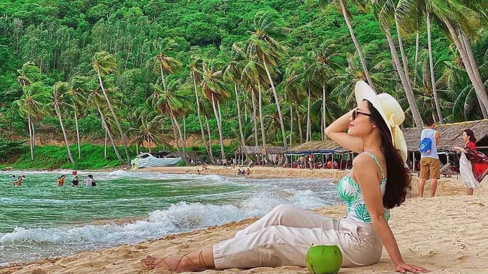 A woman sitting on a beach with palm trees and a boat in the background. 5