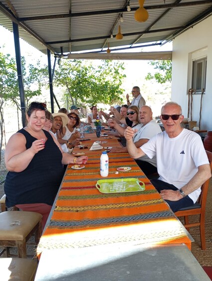 A group of people sitting around a long table outdoors under a metal roof, with a patterned tablecloth and a bottle of milk on the table. 4