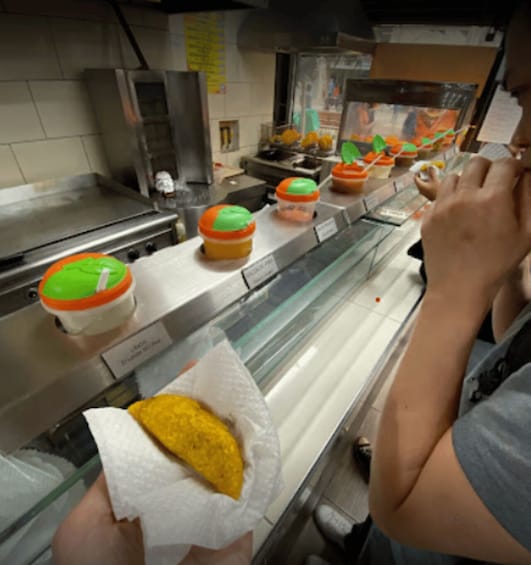 A person holding a wrapped food item, possibly a taco, in a food service area with a counter displaying various food items and containers. 3