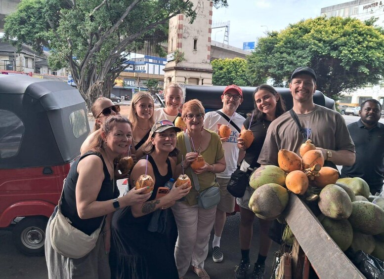 A group of people standing outdoors, some holding coconuts and others holding drinks, with a red auto-rickshaw and a fruit stand in the background. 4