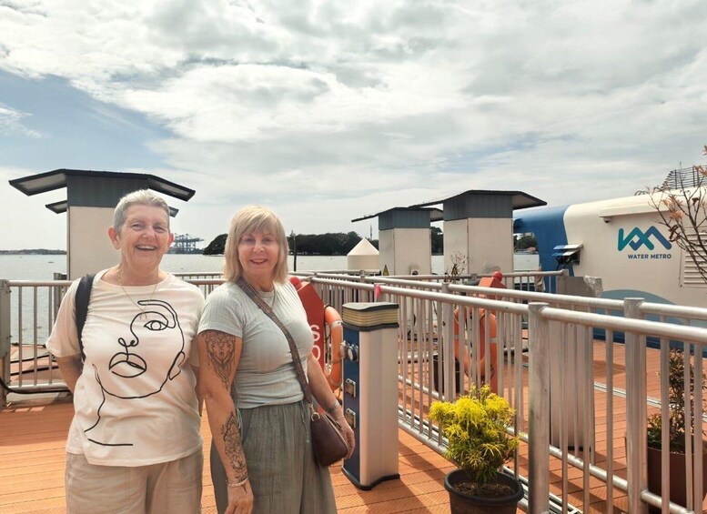 Two people standing on a deck with a water body and industrial structures in the background. 1