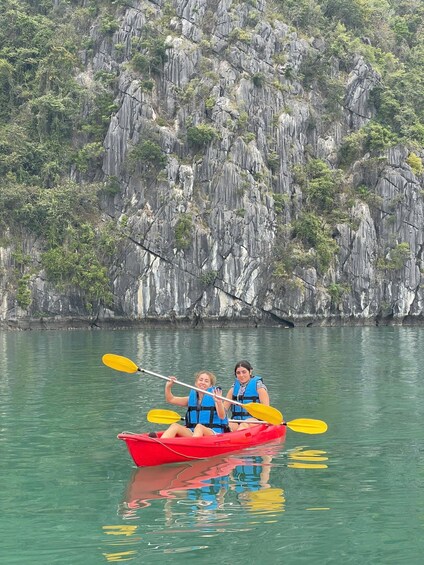 Two people kayaking in a red boat with yellow paddles on calm water, with a rocky cliff in the background. 2