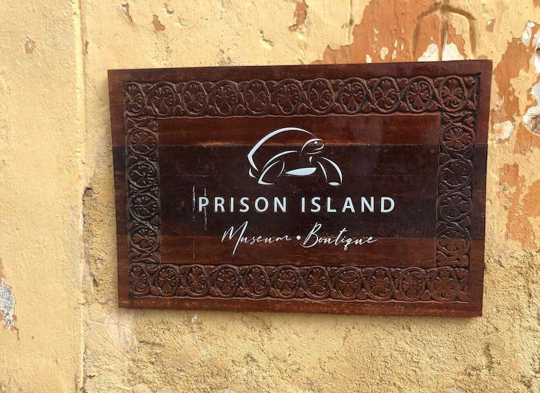 A wooden sign with a crab logo and text reading "Prison Island Museum Boutique" against a textured wall. 3
