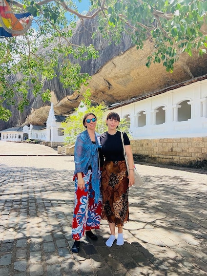 Two people standing in front of a white building with arches, with a large rock formation and trees in the background. 5