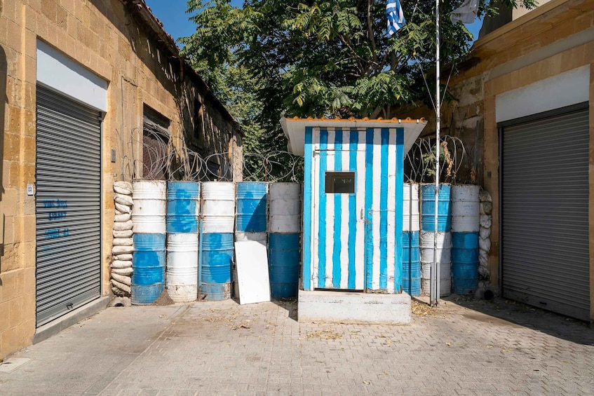 A narrow alley with closed metal shutters, blue and white barrels, and a small blue and white structure. 1