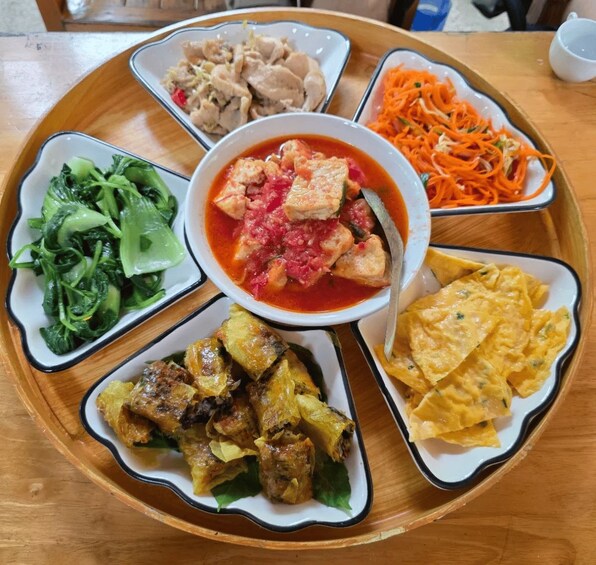 A variety of dishes on a wooden tray, including a bowl of red soup, a plate of shredded carrots, a dish of leafy greens, a plate of fried meat, and a bowl of yellow rice. 2