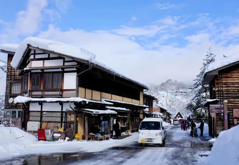 A snow-covered street with traditional wooden buildings, a car, and people in winter clothing. 5