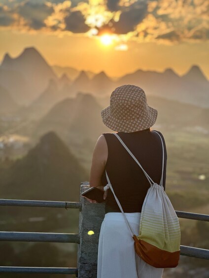 A person wearing a hat and a sleeveless top, standing on a railing with a mountainous landscape in the background. 1