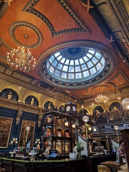 An ornate bar with a large circular skylight, chandeliers, and vintage decor. 3