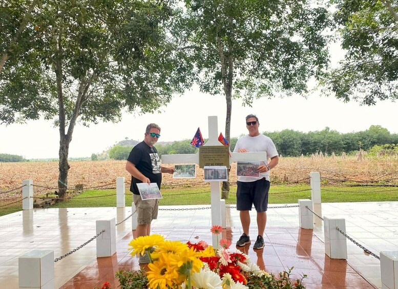 Two people at a memorial with a plaque, flowers, and flags. 1