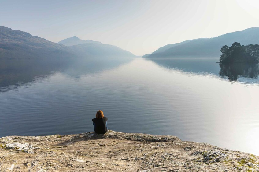 A person sitting on a rocky outcrop overlooking a calm lake with mountains in the background. 2
