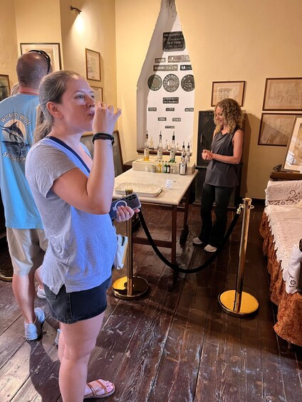 A woman drinking from a glass, standing near a table with bottles and glasses, in a room with framed pictures on the wall. 2